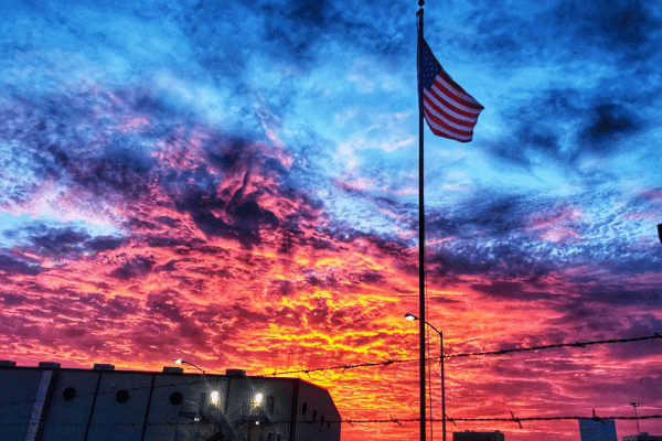Image of a United States flag with a building in the background with the sun going down on a cloudy day.
