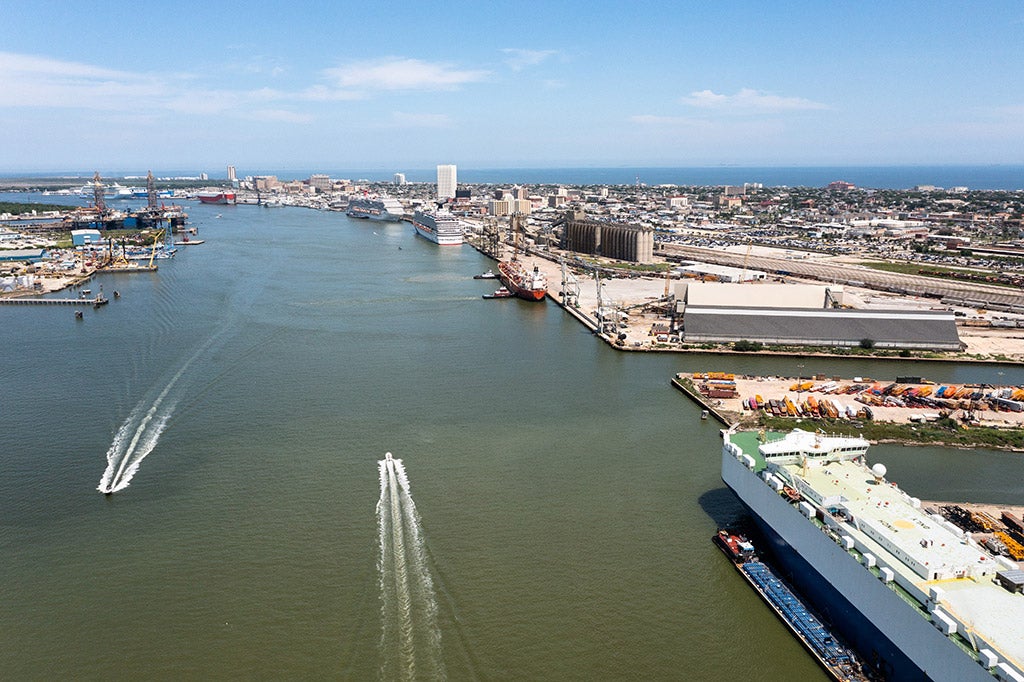 Aerial view of the Galveston Harbor looking west, showing port facilities, docks, cargo areas, and the surrounding waterfront, with the harbor waters and skyline in the distance.